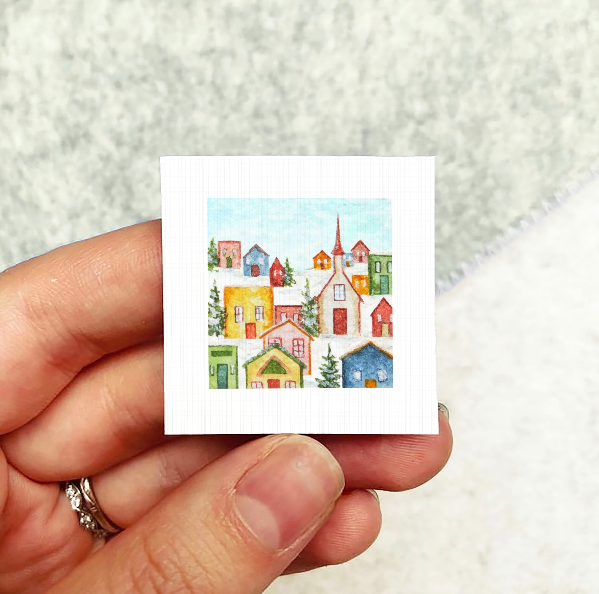 A hand holding a small square-shaped paper with a colorful illustration of a winter village scene.