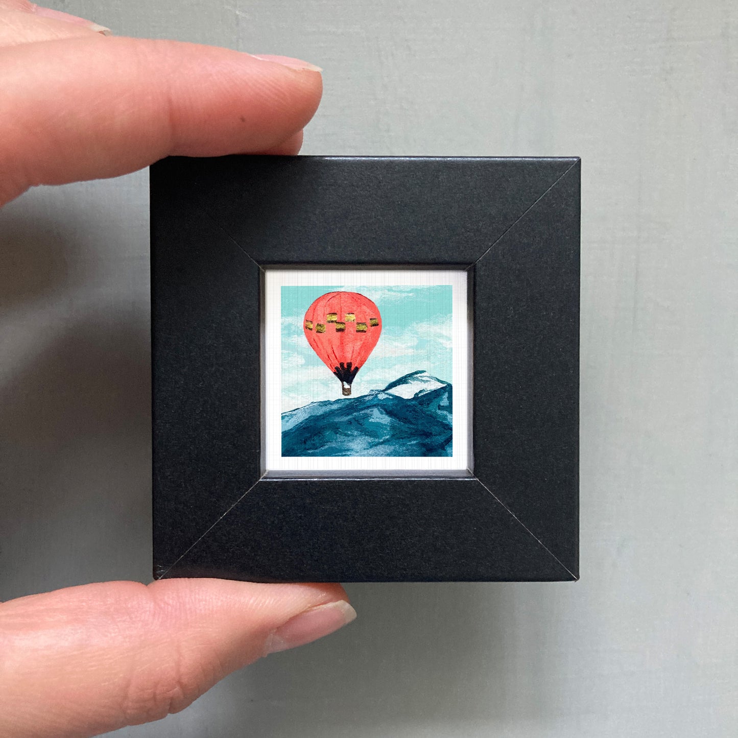 A hand is holding a small black frame containing a framed image of a hot air balloon floating over a mountain landscape.