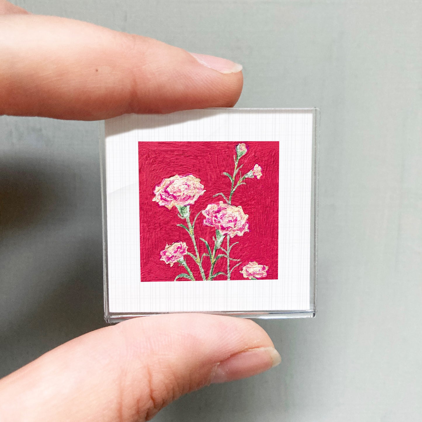 A person's hand is holding a small square picture with a vibrant red background and delicate pink flowers.
