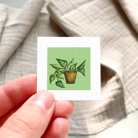 A hand is holding a small square card with a painting of a potted plant on it.