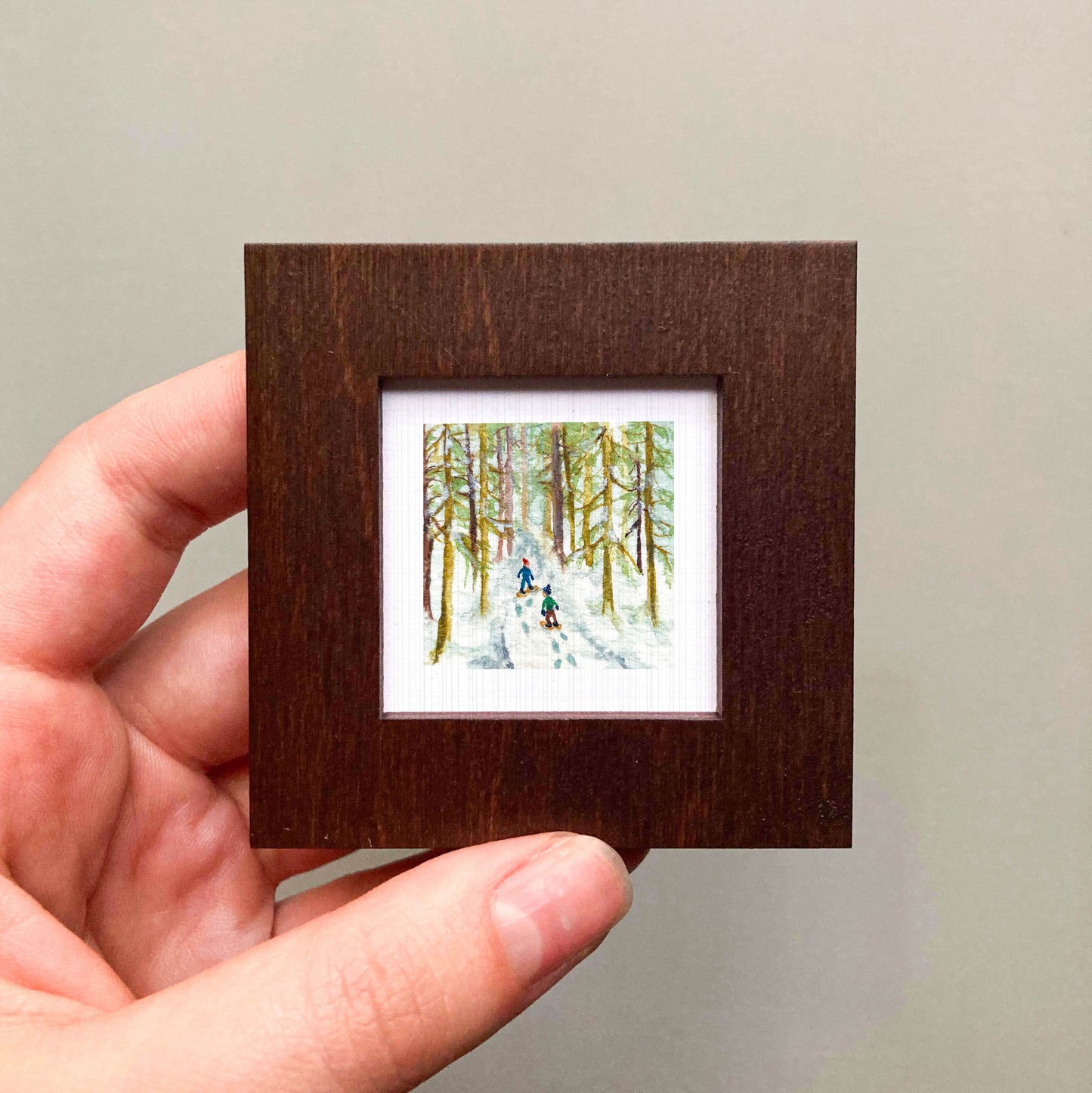 A hand is holding a small, square-shaped wooden frame containing a painting of two people skiing in a snowy forest.
