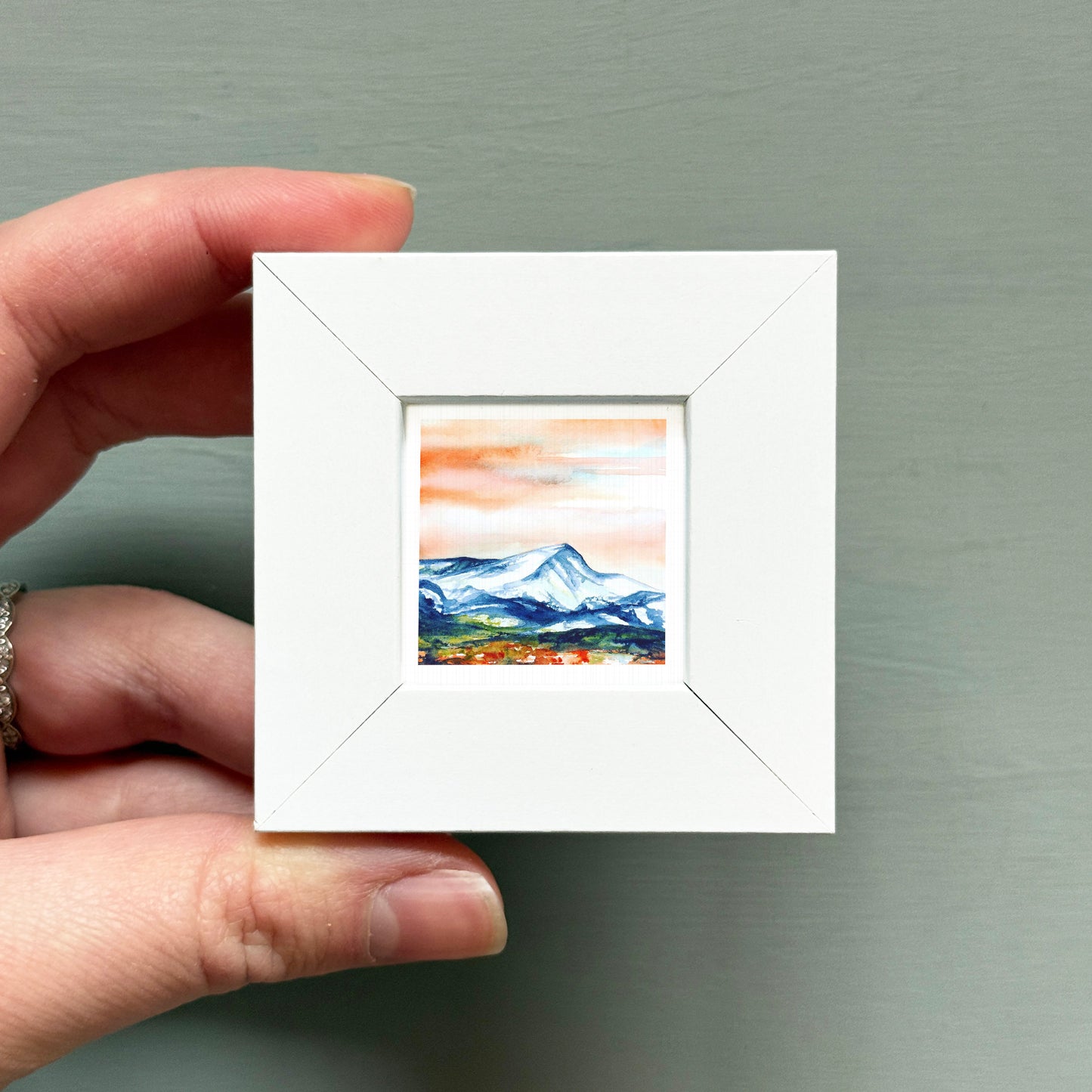 A hand is holding a small white square frame containing a painting of a mountain landscape.