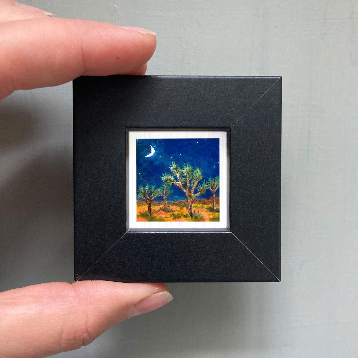 A hand is holding a small black frame containing a framed painting of a desert landscape at night, with a crescent moon and stars in the sky.
