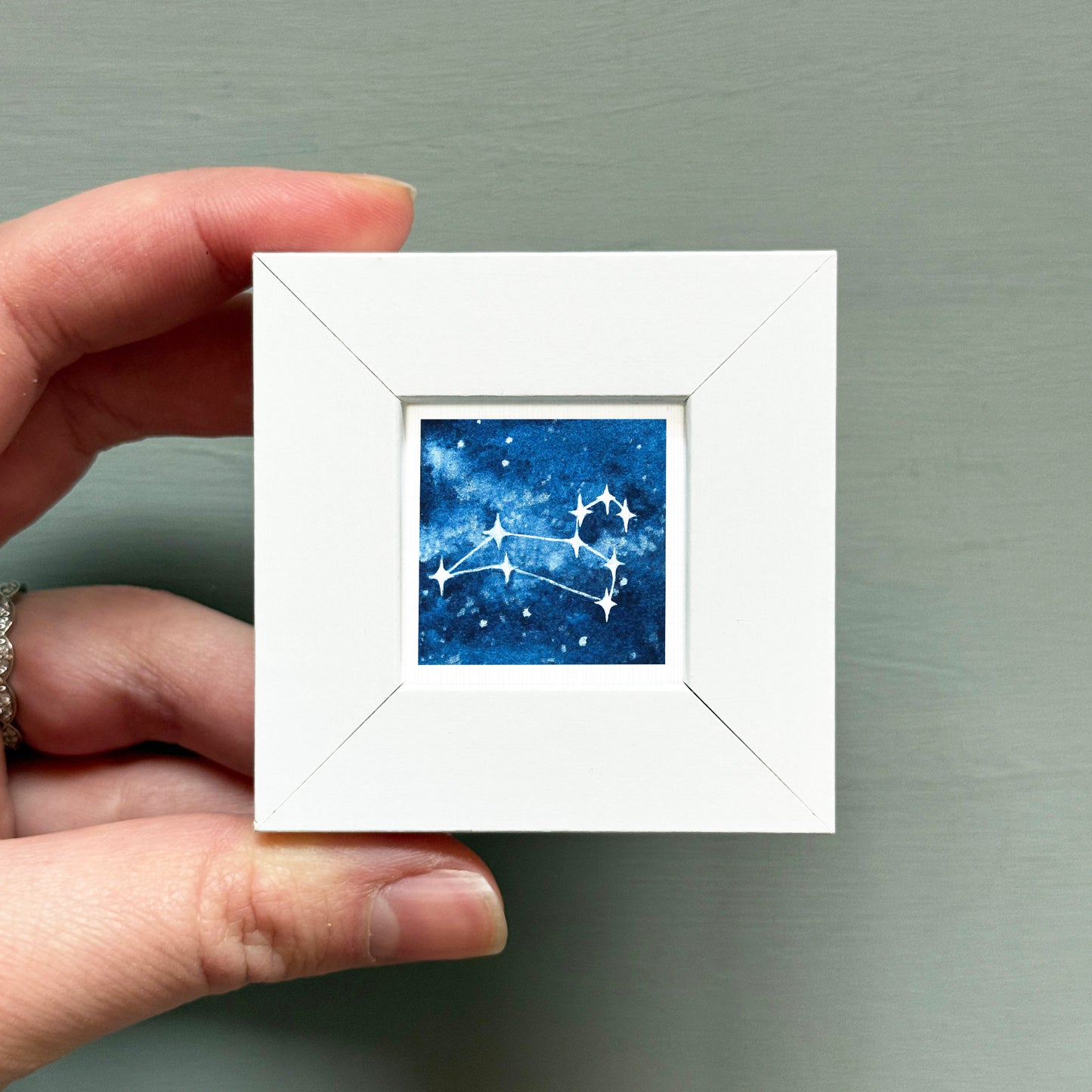 A hand is holding a small white square frame containing a watercolor painting of a starry night sky.