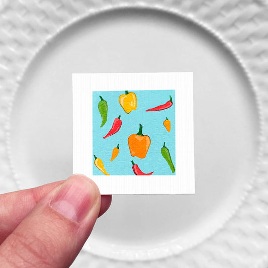 A hand is holding a small square with a vibrant illustration of various colorful peppers on a blue background.