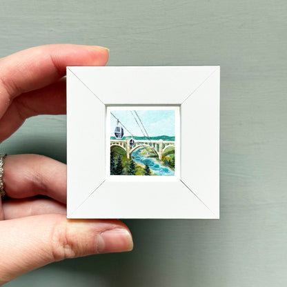 A hand is holding a small white square frame containing a painting of a bridge and a gondola.
