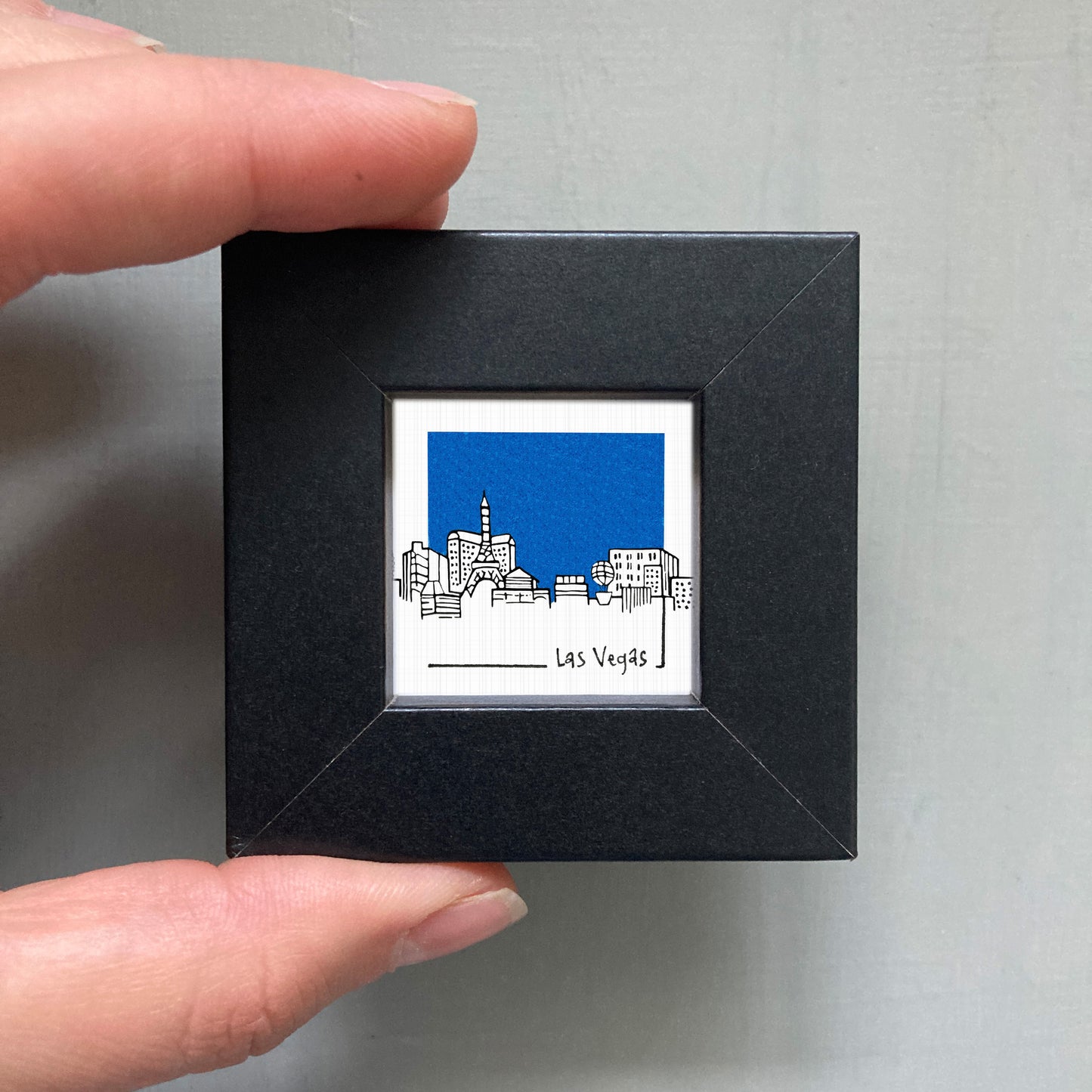 A person's hand holding a small square frame containing an illustration of a city skyline with a blue sky.
