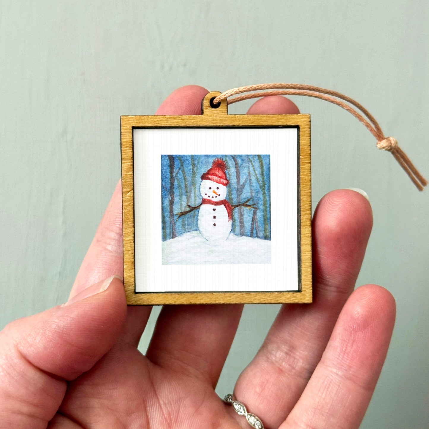 A hand holding a small wooden frame containing a picture of a snowman.