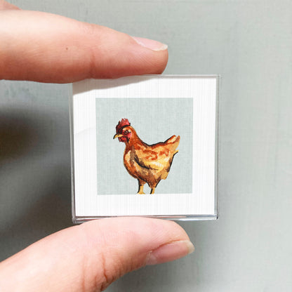 A person's hand is holding a small square picture of a chicken.