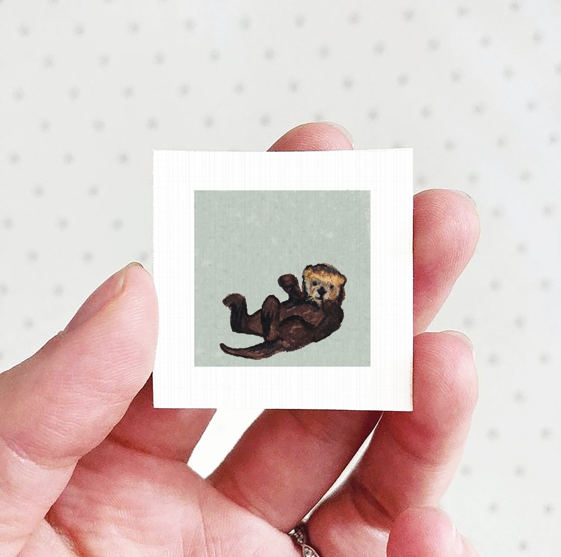 A person's hand holding a small square picture of an otter.