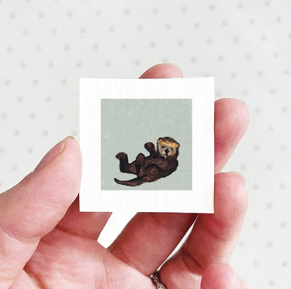 A person's hand holding a small square picture of an otter.