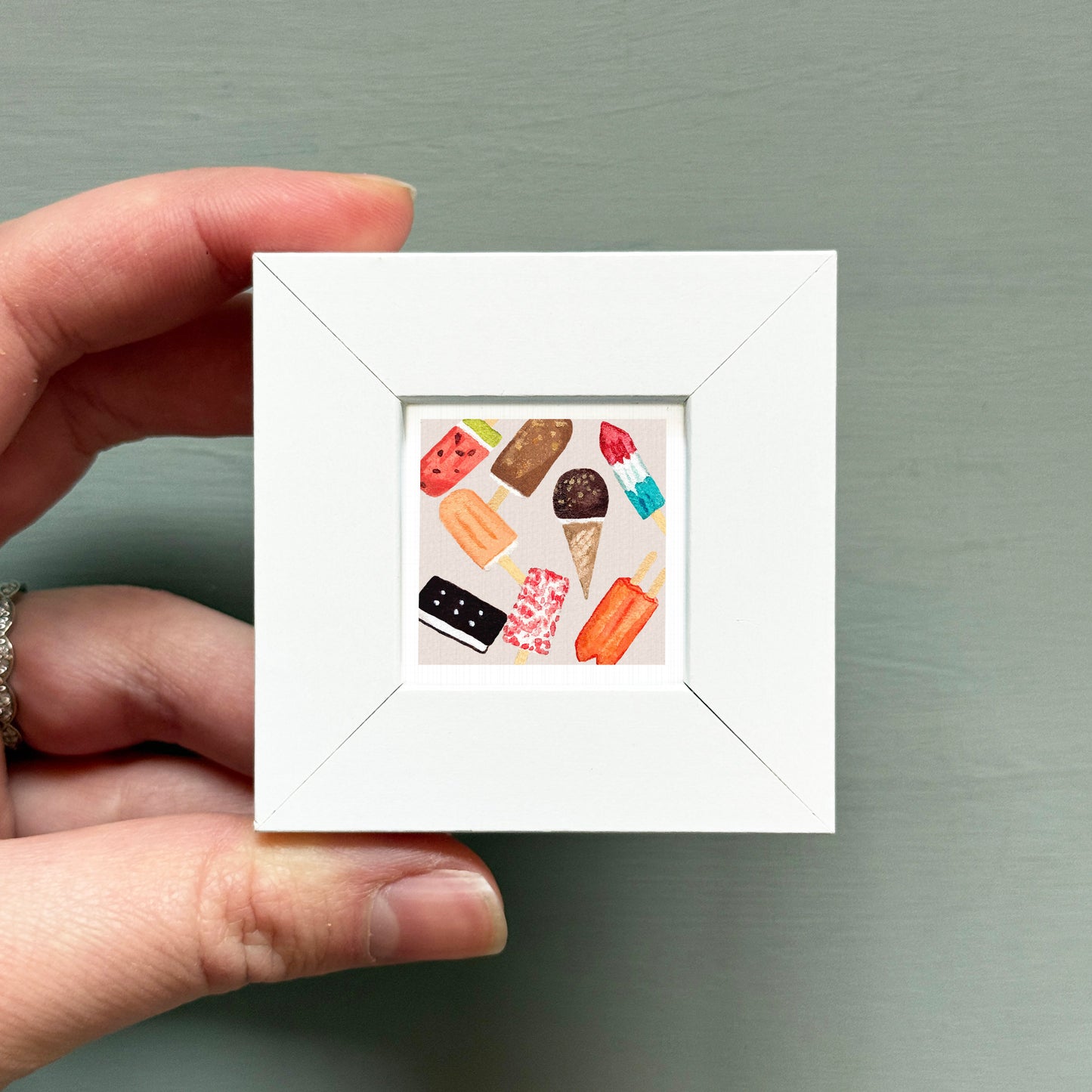 A hand is holding a small white square frame containing an illustration of various colorful popsicles.
