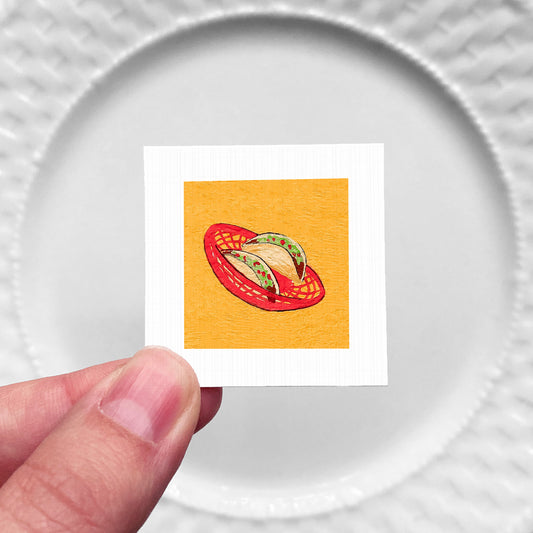 A hand is holding a small square with an illustration of a red basket containing two tacos on a yellow background.