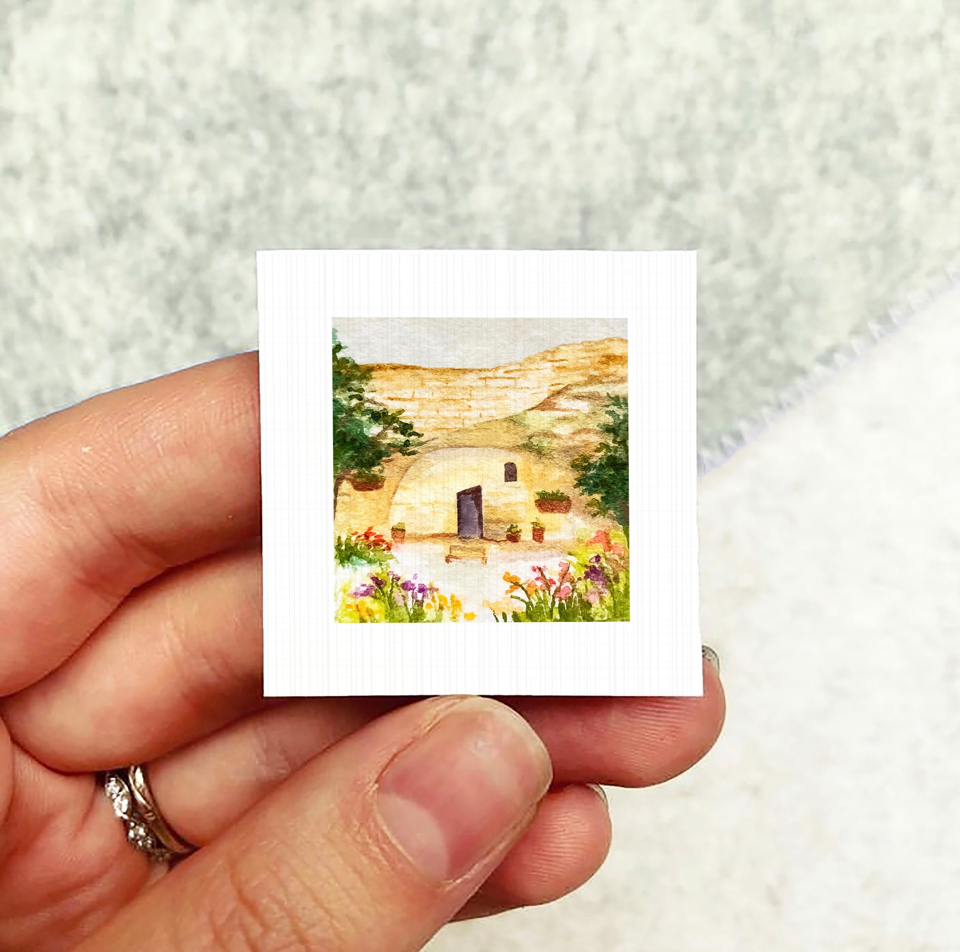 A hand holding a small square picture depicting a house surrounded by flowers and greenery.