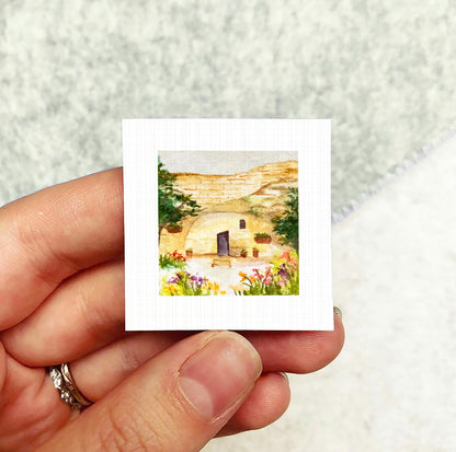A hand holding a small square picture depicting a house surrounded by flowers and greenery.