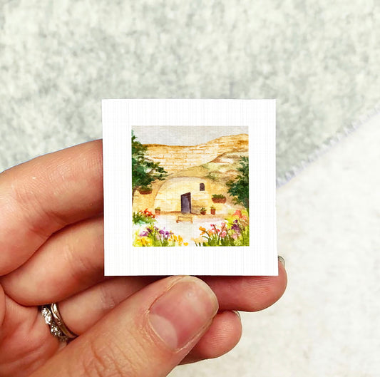 A hand holding a small square picture depicting a house surrounded by flowers and greenery.