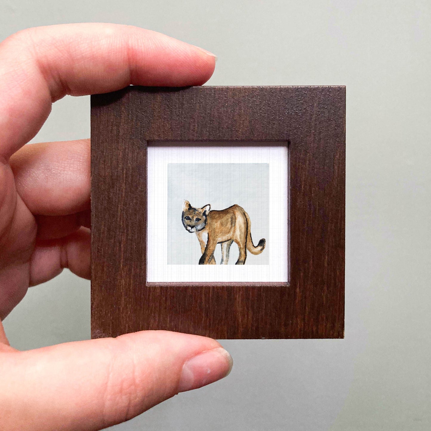 A hand is holding a small wooden frame containing a painting of a brown mountain lion.
