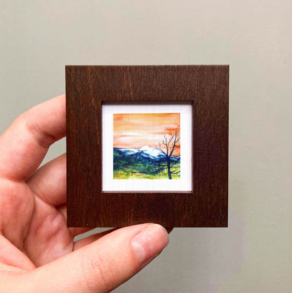 A hand is holding a small, square-shaped wooden frame containing a painting of a mountain landscape with a tree in the foreground.