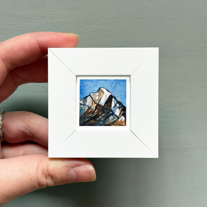 A hand is holding a small white square frame containing a painting of a mountain landscape.
