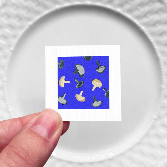 A hand is holding a small square with a pattern of mushrooms against a blue background.