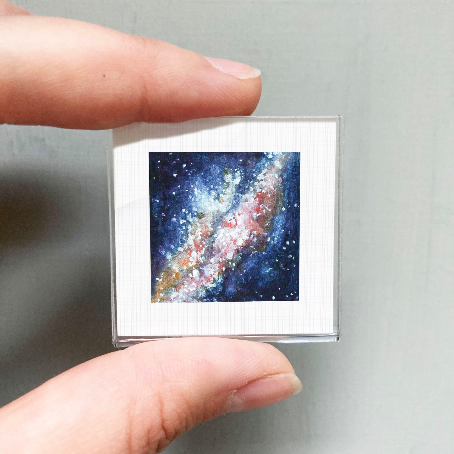 A person's hand holding a small square picture of a colorful galaxy against a gray background.