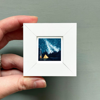 A hand is holding a small white square frame containing a painting of a starry night sky with a tent in the foreground.