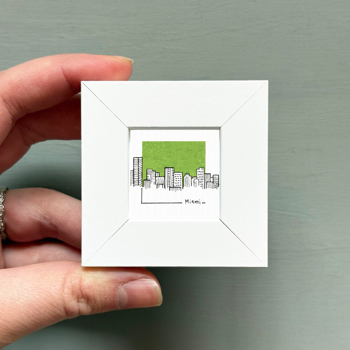 A hand is holding a small white square frame containing a miniature illustration of a city skyline.