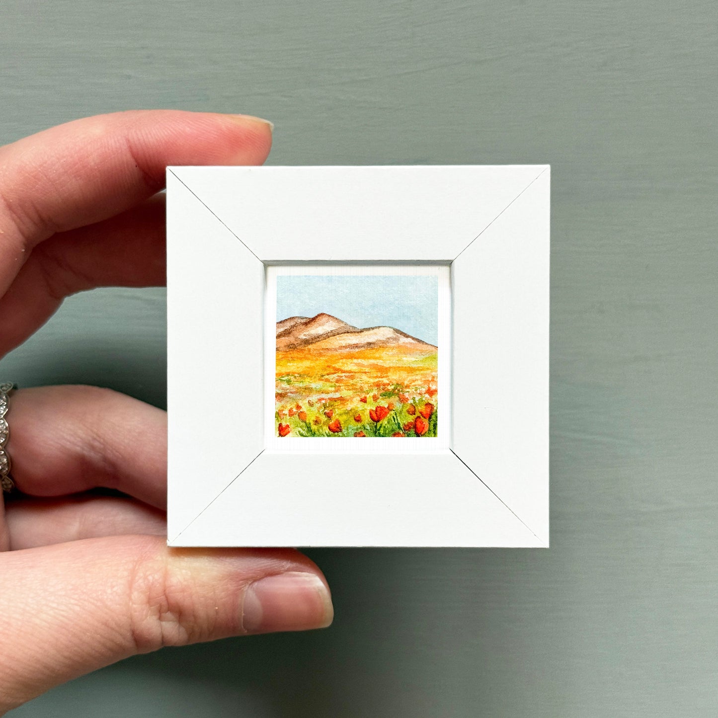 A hand is holding a small white picture frame containing a painting of a field with mountains in the background.