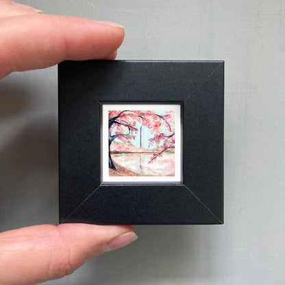 A person's hand holding a small black picture frame containing a framed painting of a cherry blossom tree.