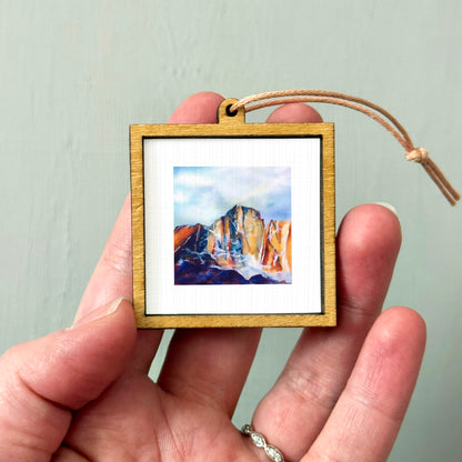 A hand holding a small wooden frame containing a watercolor painting of a mountain landscape.