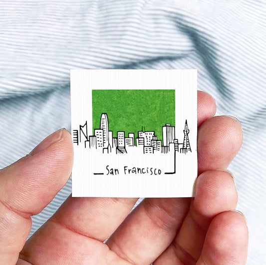 A hand holding a small square-shaped object with a green background and a cityscape illustration on it.