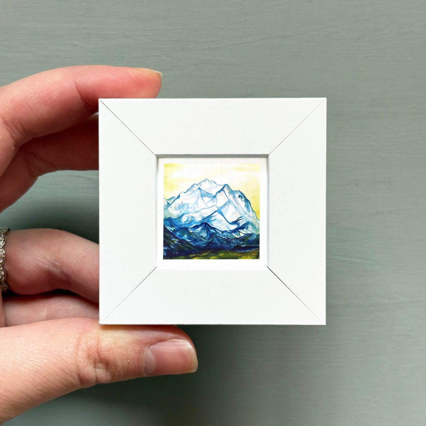 A hand is holding a small white square frame containing a painting of a mountain landscape.