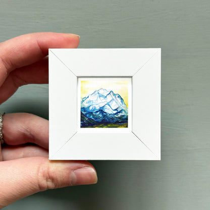 A hand is holding a small white square frame containing a painting of a mountain landscape.