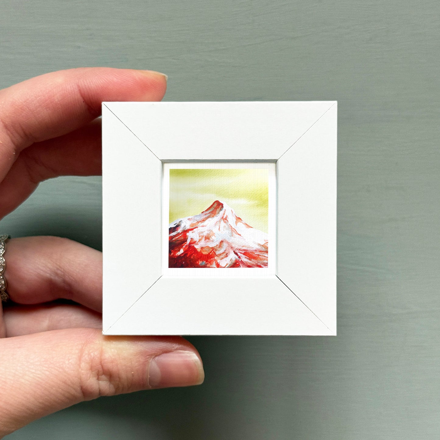A hand is holding a small white square frame containing a painting of a mountain landscape.