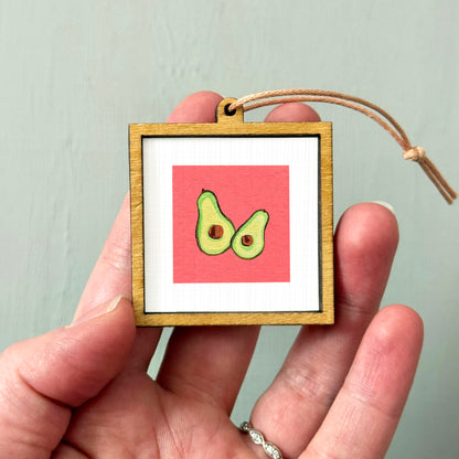 A hand holding a small wooden frame with a picture of two avocados inside it.