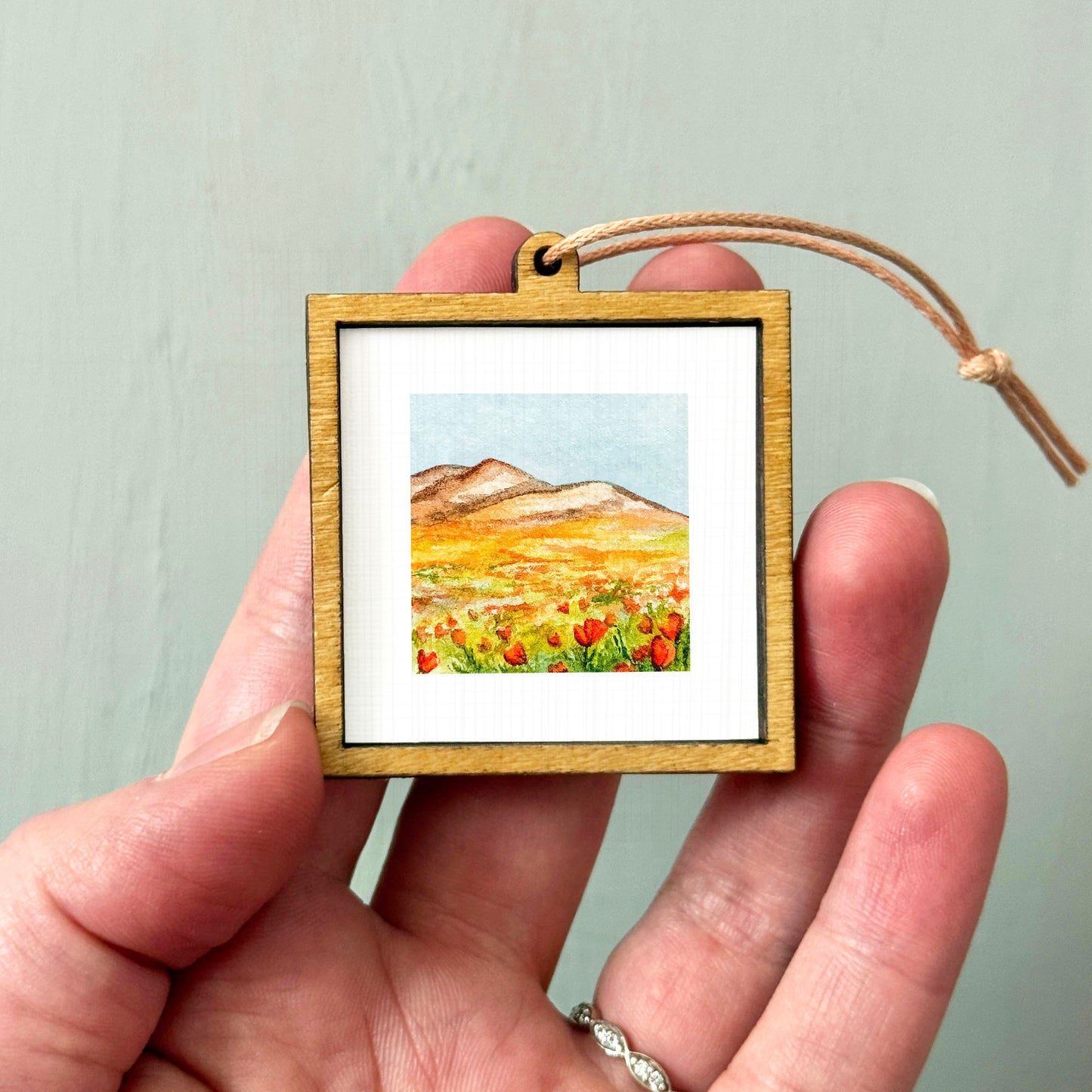 A hand holding a small wooden frame containing a watercolor painting of a mountain landscape with a field of flowers.