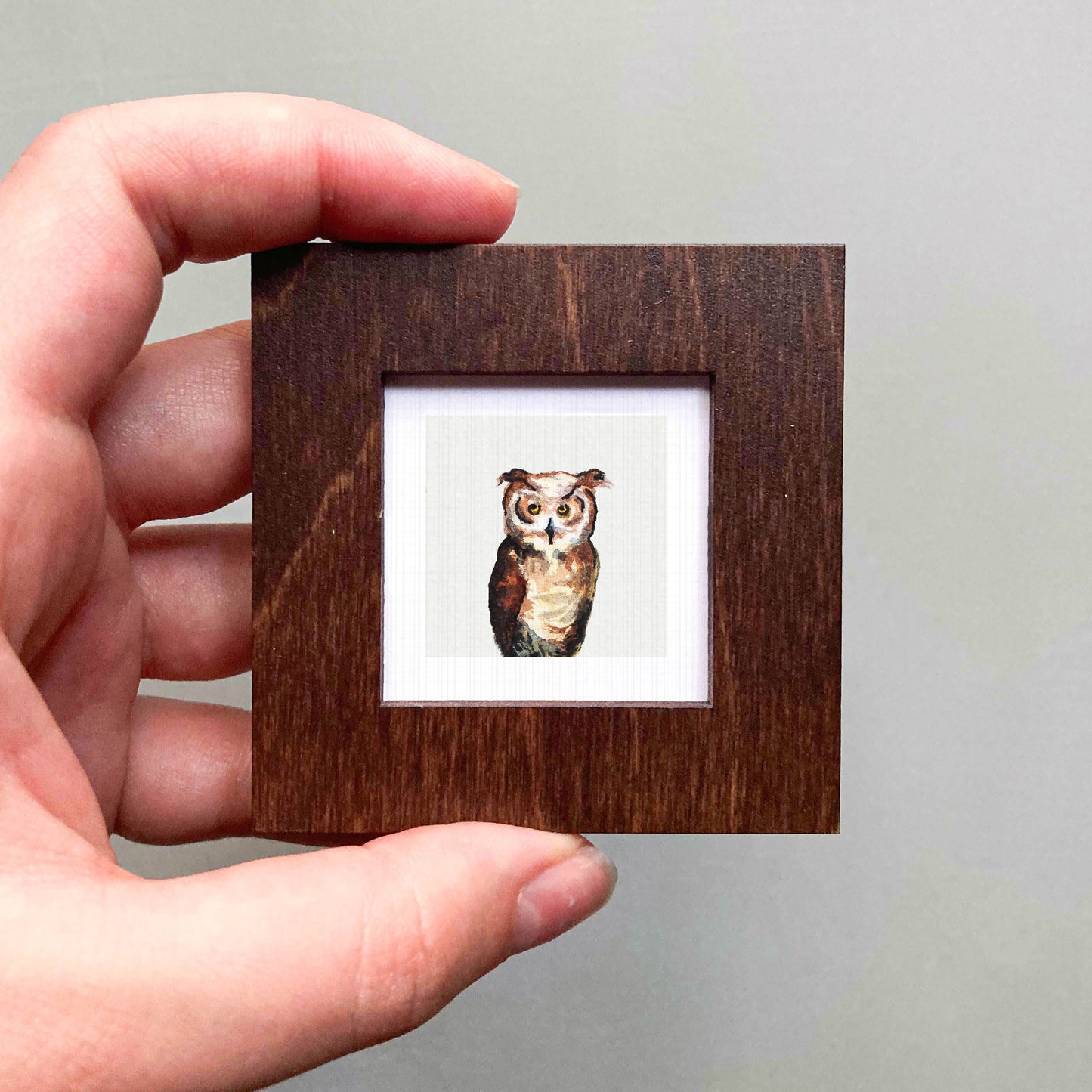 A hand is holding a small wooden frame containing a picture of an owl.