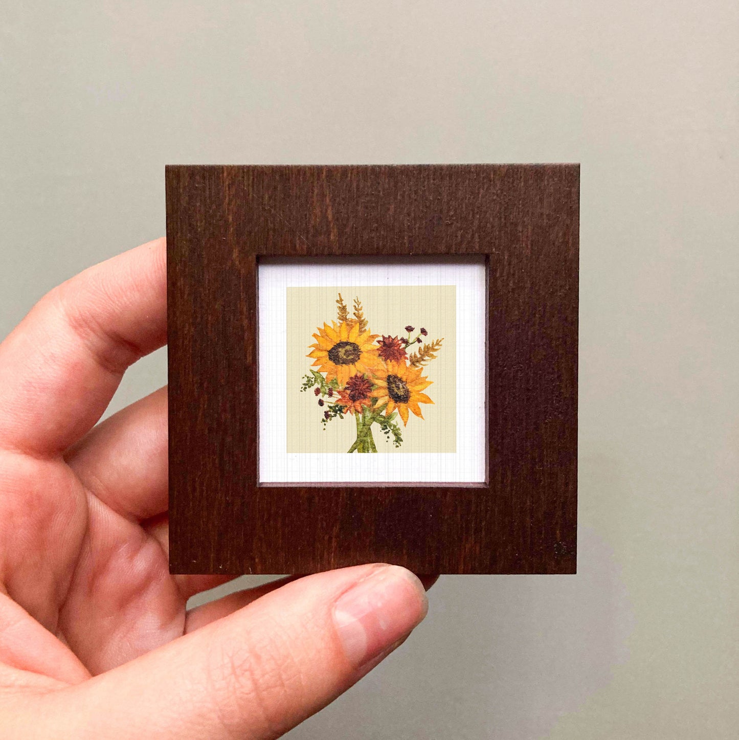 A hand is holding a small wooden frame containing a framed picture of a bouquet of sunflowers.