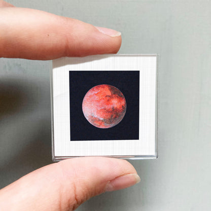 A person's hand is holding a small square frame containing a photograph of a red planet, likely the moon, against a dark background.
