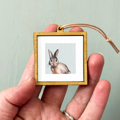 A person's hand holding a small wooden frame containing a picture of a rabbit.