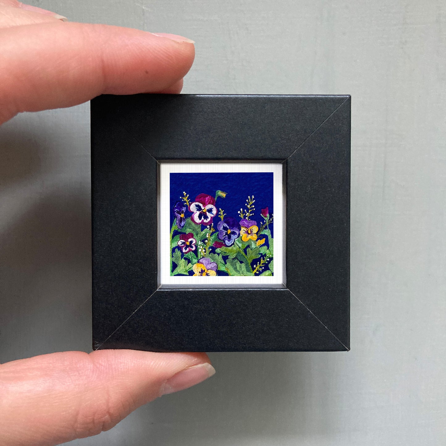 A hand is holding a small black picture frame containing a vibrant painting of colorful flowers.