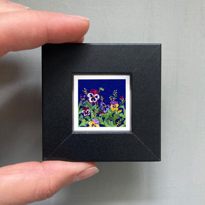 A hand is holding a small black picture frame containing a vibrant painting of colorful flowers.
