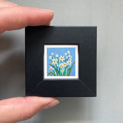 A person's hand holding a small square picture frame containing a painting of white flowers against a blue background.