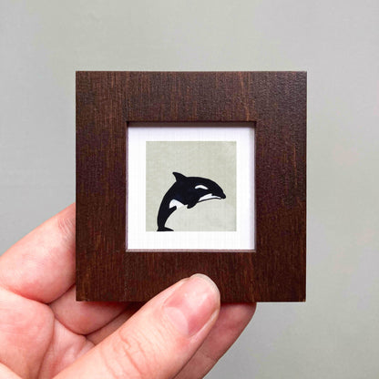 A hand is holding a small wooden frame containing a black and white image of a whale.