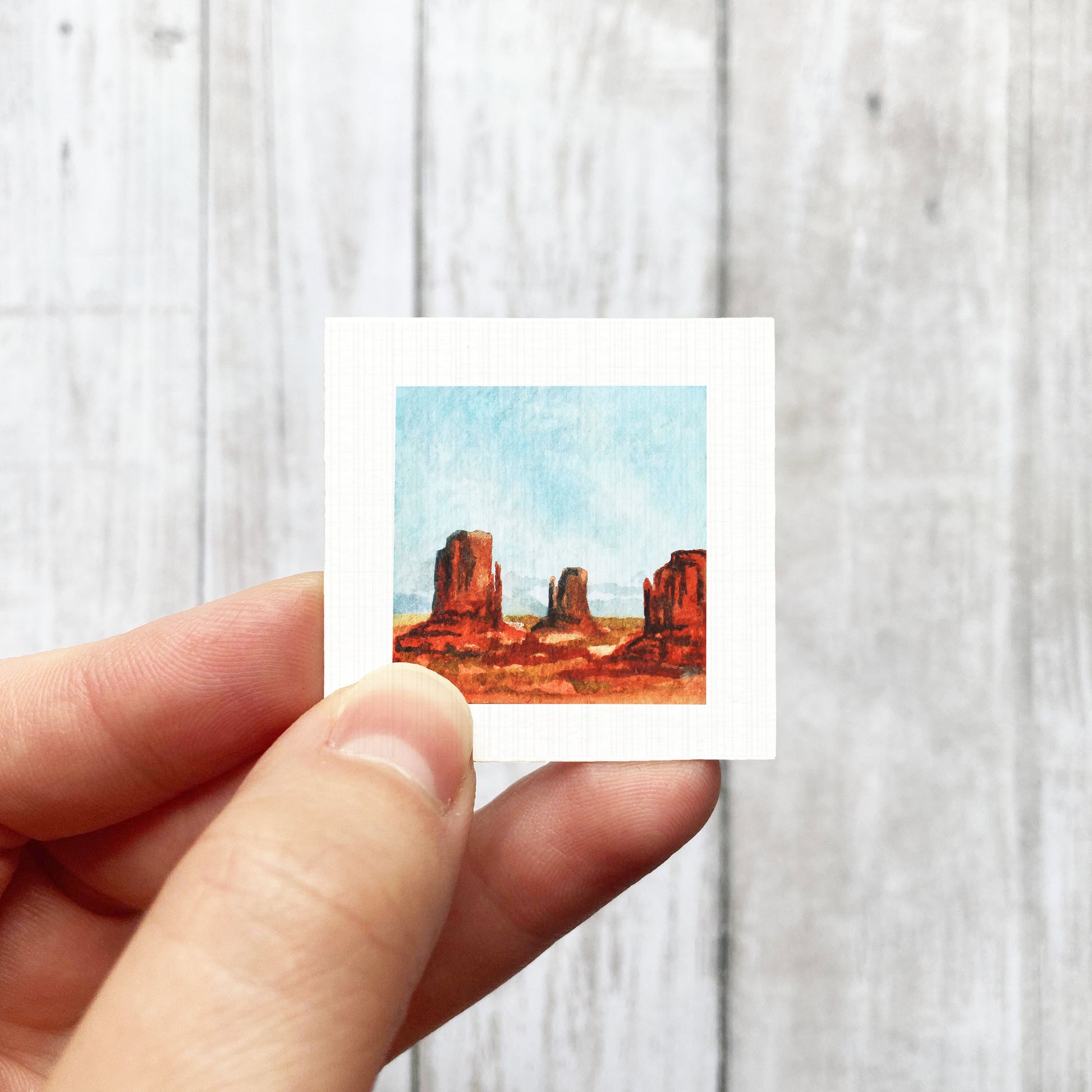 A hand is holding a small square-shaped painting depicting a desert landscape with red rock formations.