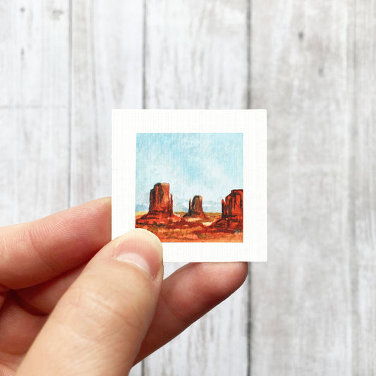 A hand is holding a small square-shaped painting depicting a desert landscape with red rock formations.