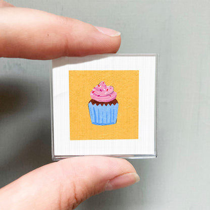 A hand holding a small square card with a colorful illustration of a cupcake on it.