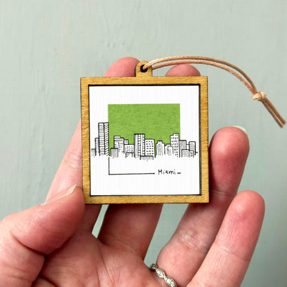 A hand holding a small wooden frame with a picture of a city skyline inside it.