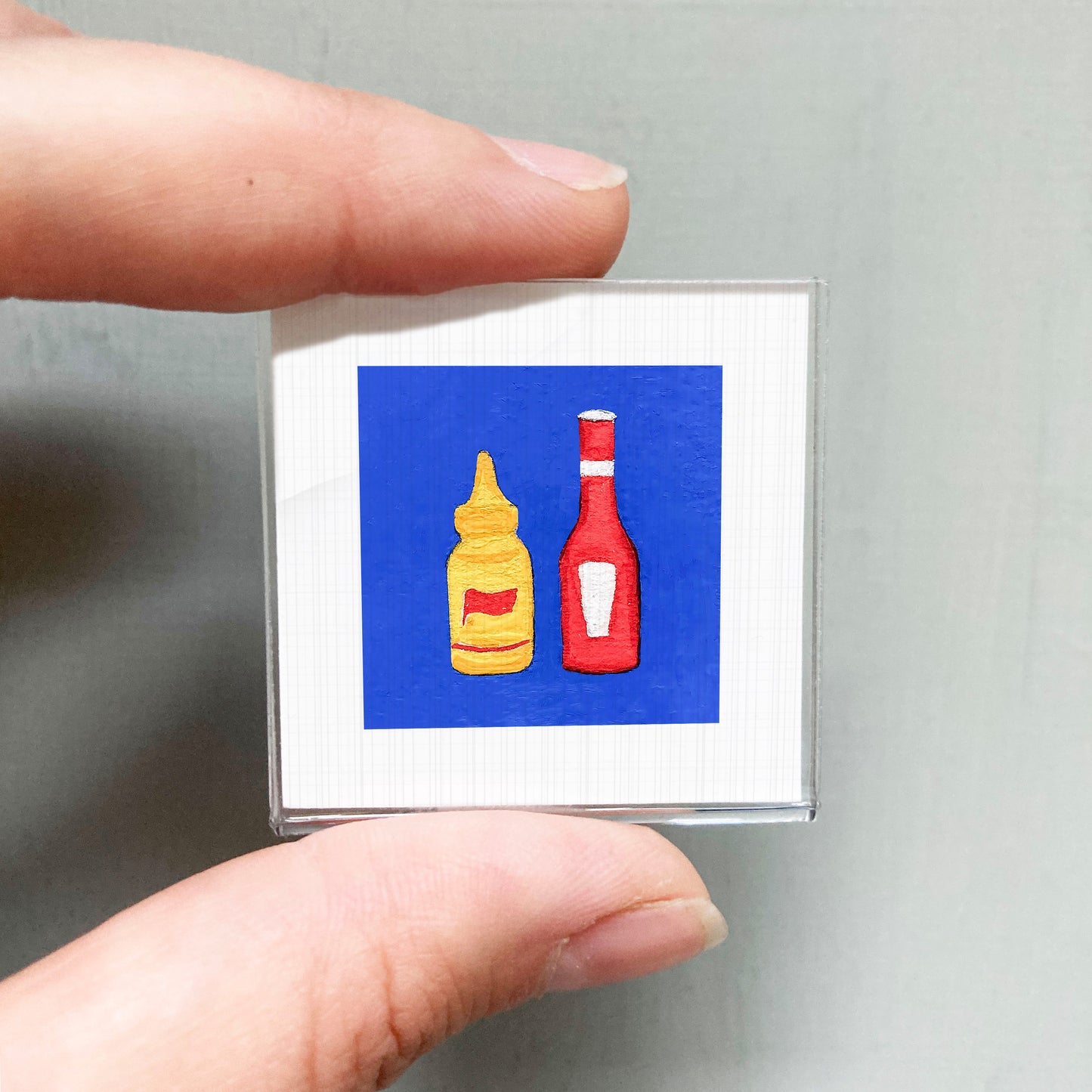 A person's hand holding a small square with two bottles of condiments, one yellow and one red, against a blue background.