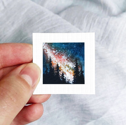 A hand is holding a small square-shaped piece of paper with a painting of a night sky with stars and trees on it.